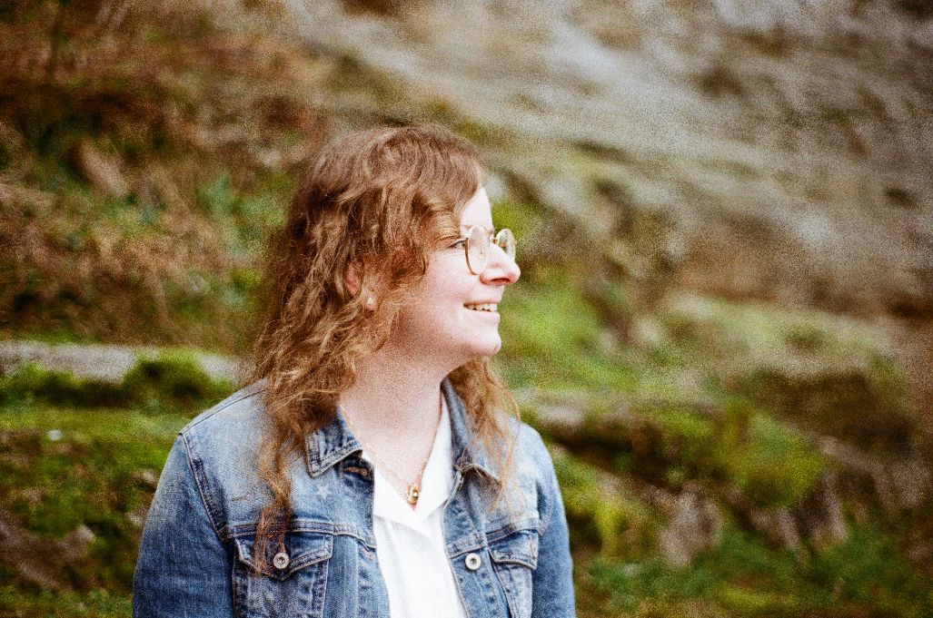 Rahel in Jeansjacke nur mit Oberkörper von der Seite, lächelnd, vor mosigen Felswand als Hintergrund