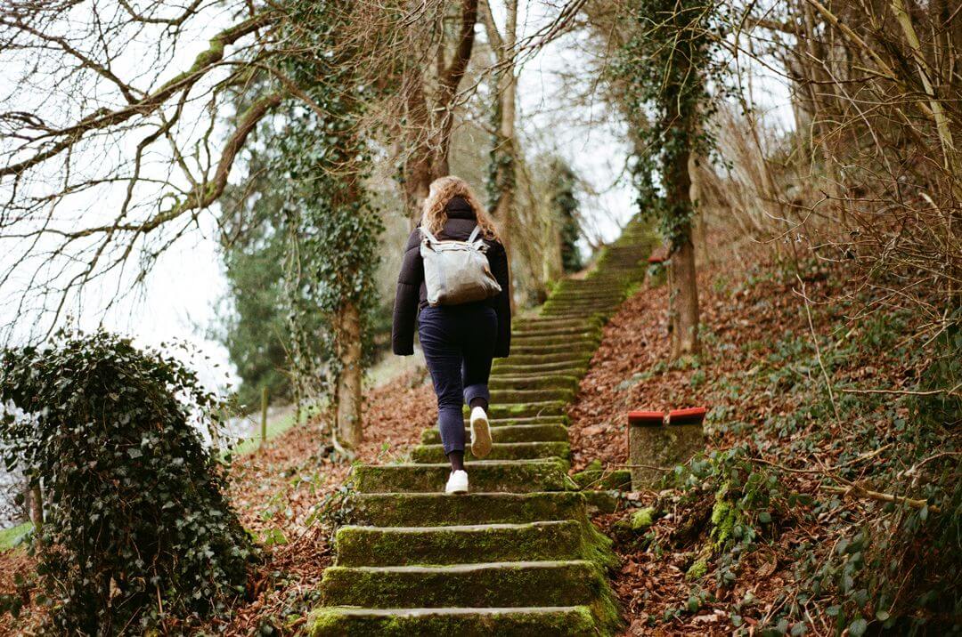 mosig grüne Steintreppe nach oben im Februar. Gesäumt von braunem Laub und Bäumen mit Efeu links und rechts. Rahel ganzkörper und von hinten abgebildet, geht die Treppe hoch. In Winterjacke, blauer Hose und mit grauem Rucksack.